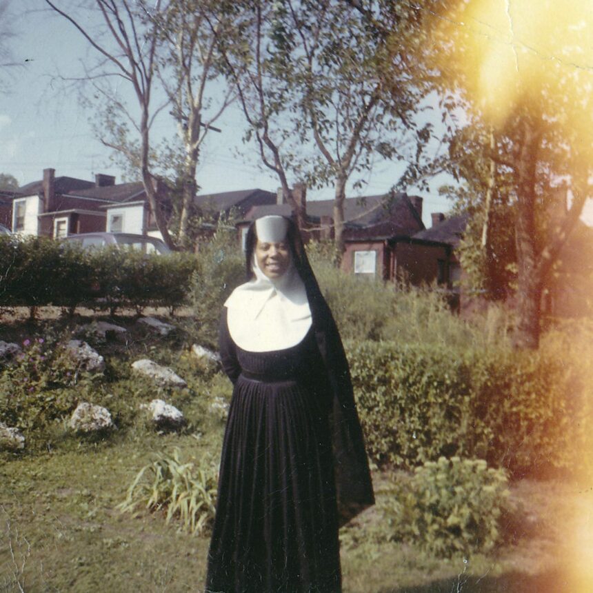 A nun in black dress standing outside near bushes.