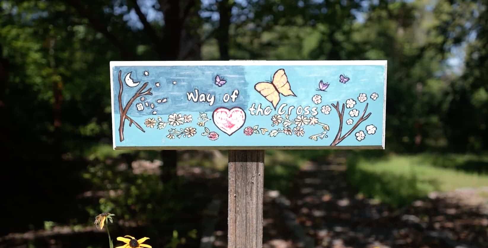 A blue way of the cross sign with trees, butterflies, and hearts painted on it.