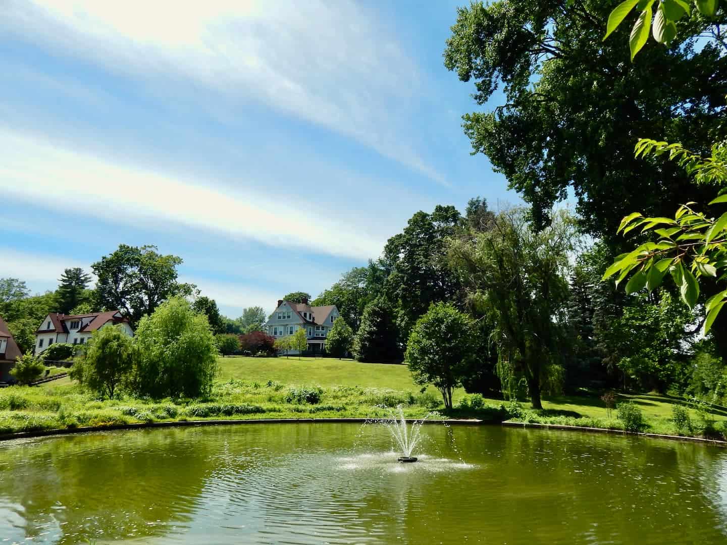 A view of Cranaleith with the fountain in the front surrounded by trees, the historic house, and administration building. The sky is a beautiful mix of blue and white.