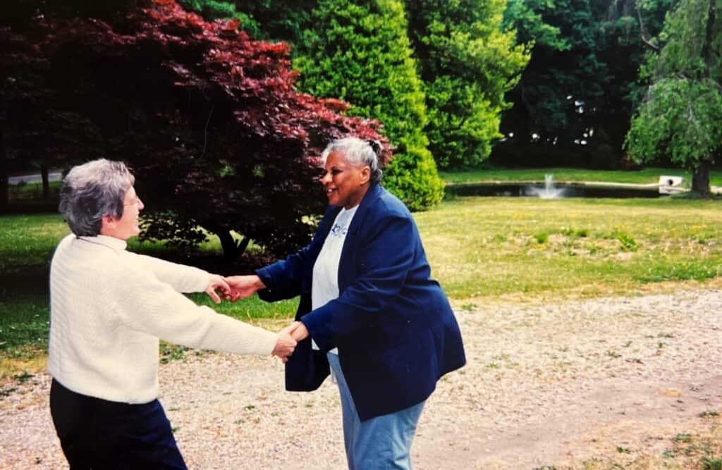 Sister Maria holding hands with a lady with trees and the fountain in the background.