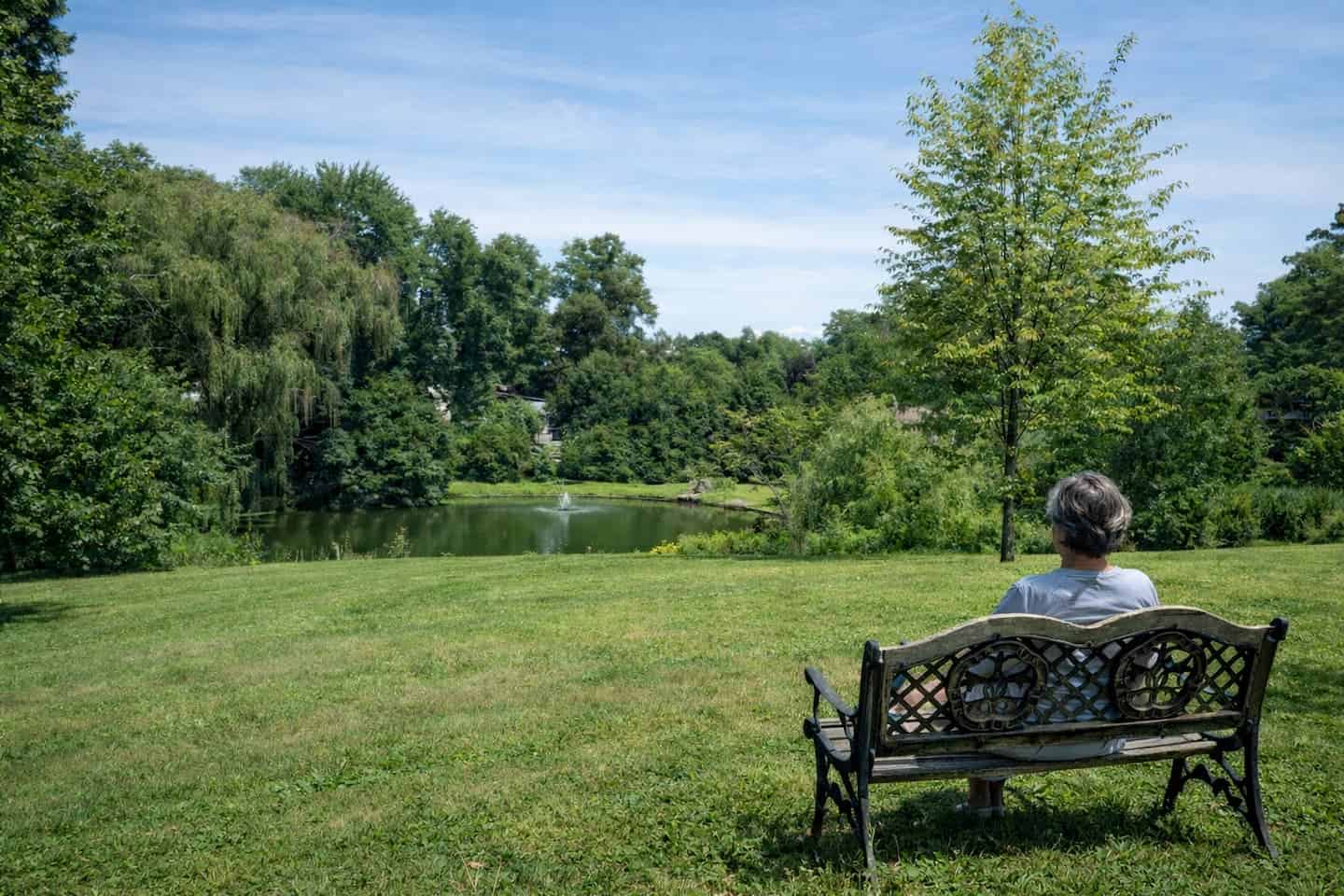 A lady sitting on a bench staring into a beautiful field and fountain.